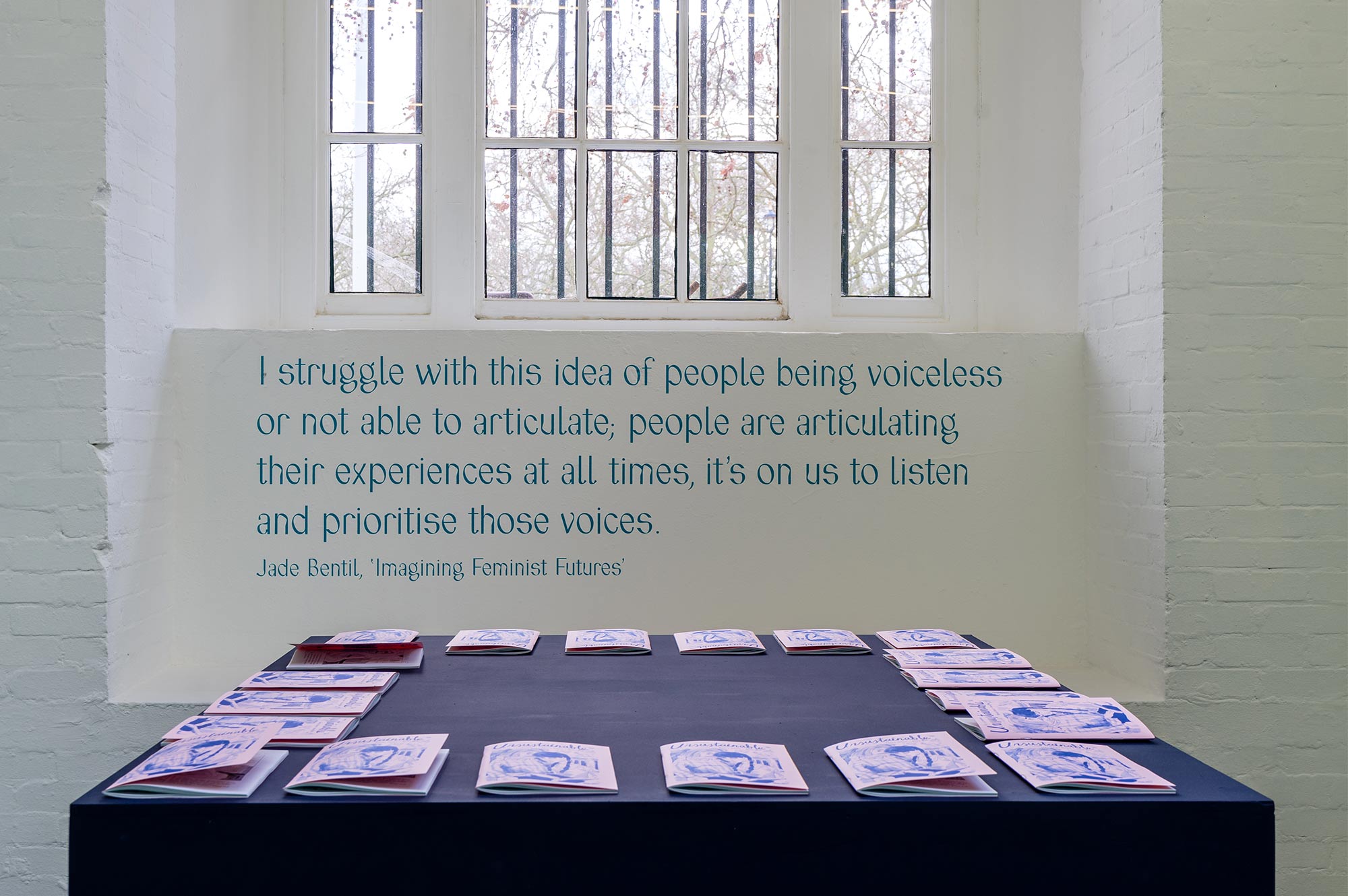 A table with pink comic books. A printed quote on the wall reads: I striggle with this idea of people being voiceless or not able to articulate; people are articulating their experiences at all times, it's on us to listen and prioritise those voices. Jade Bentil, Imagining Feminist Futures