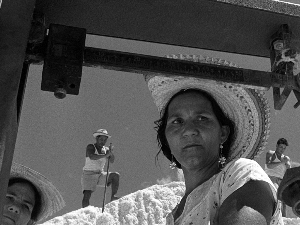 A greyscale photo. A woman looks carefully at a ruler as men work on a pile of white rocks in the background