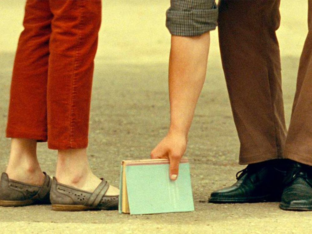 Two people stand on soil, only legs visible. One is picking up a book on the floor