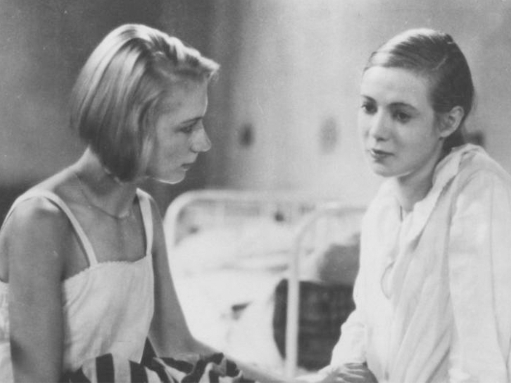 Two women sit on the side of a bed, one looking sad, the other looking on with concern