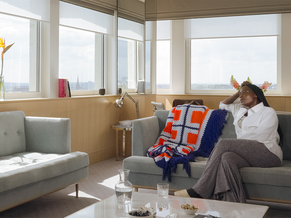 A black woman with long hair sits in a well-furnished estate apartment, looking out to a grey London sky