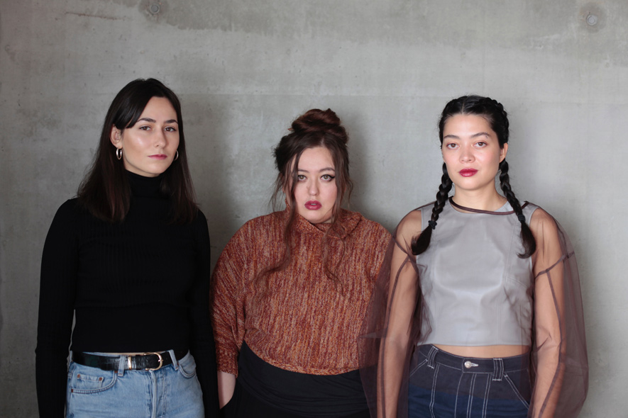 The three members of Keiken stand side by side against a grey concrete wall